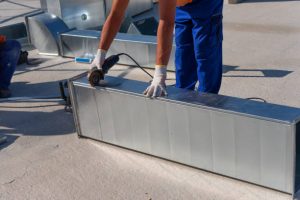 istockphoto-1191025187-612×612 HVAC technician is working on a roof of new industrial building. Close-up view of the young technician repairing an air duct with the angle grinder. HVAC worker cutting ductwork with an angle grinder.